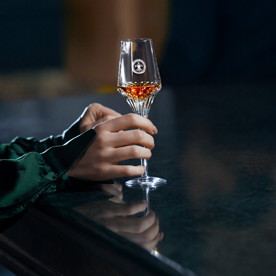 A lifestyle photo of two hands with a LOUIS XIII crystal glass, piano in the background