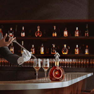 A lifestyle image of a bartender serving LOUIS XIII to a two crystal glases, a decanter on the bar counter