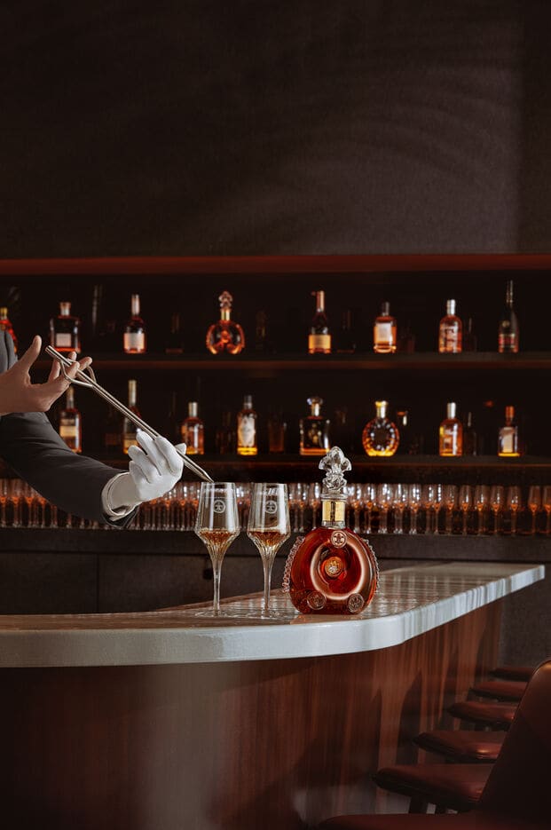 A lifestyle image of a bartender serving LOUIS XIII to a two crystal glases, a decanter on the bar counter
