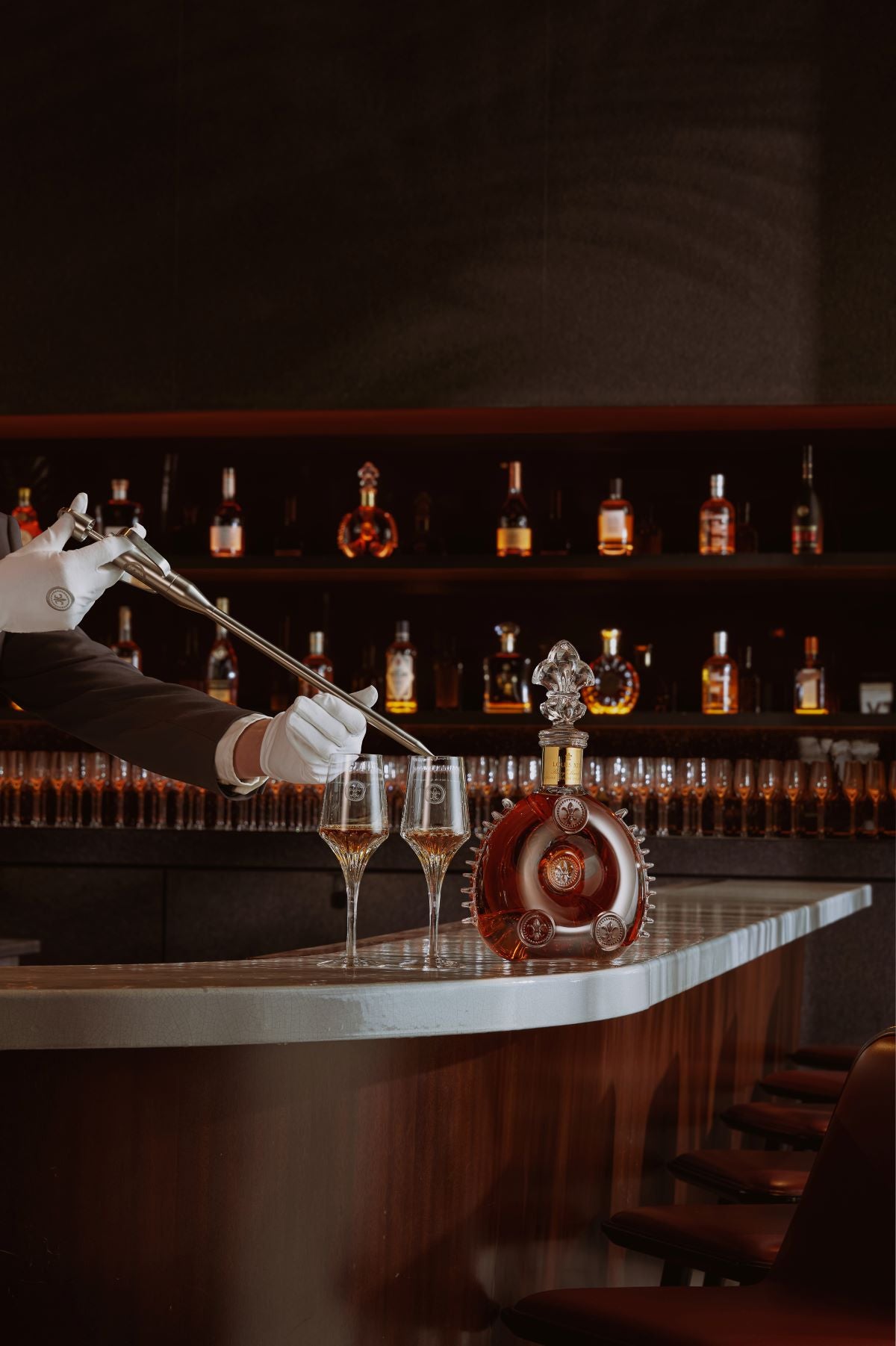 A lifestyle photo of a person with a white glove serving LOUIS XIII cognac to two crystal glasses with a spear