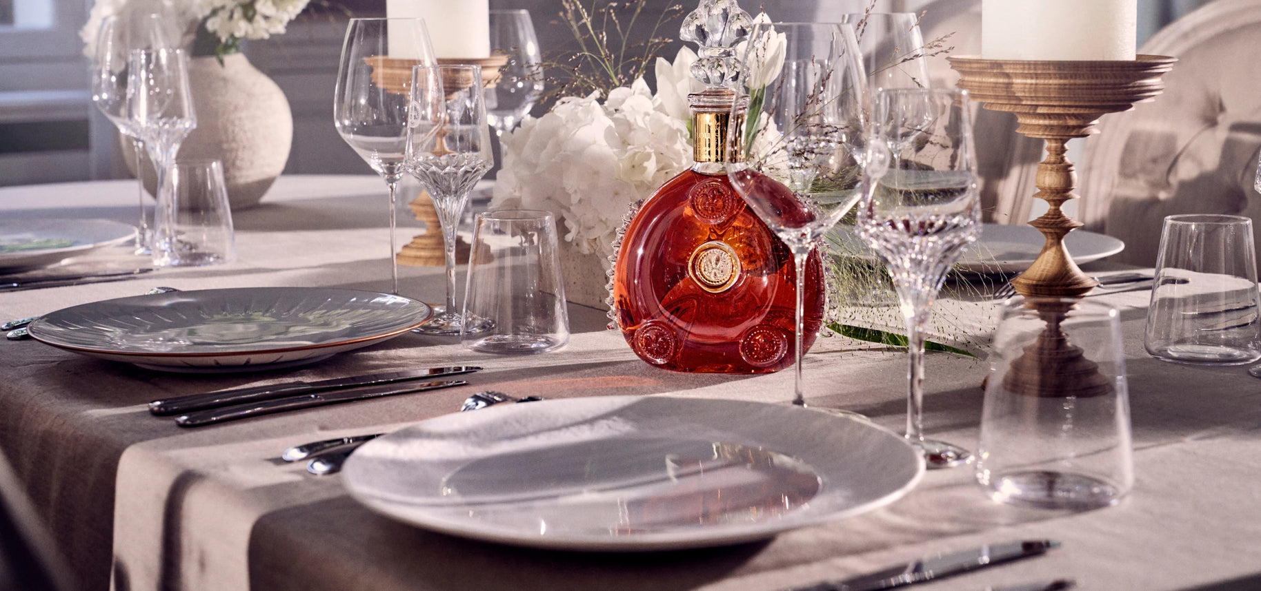 Bottle of Louis XIII cognac placed at the center of a festive arrangement with candles, flowers, plates and crystal glasses.