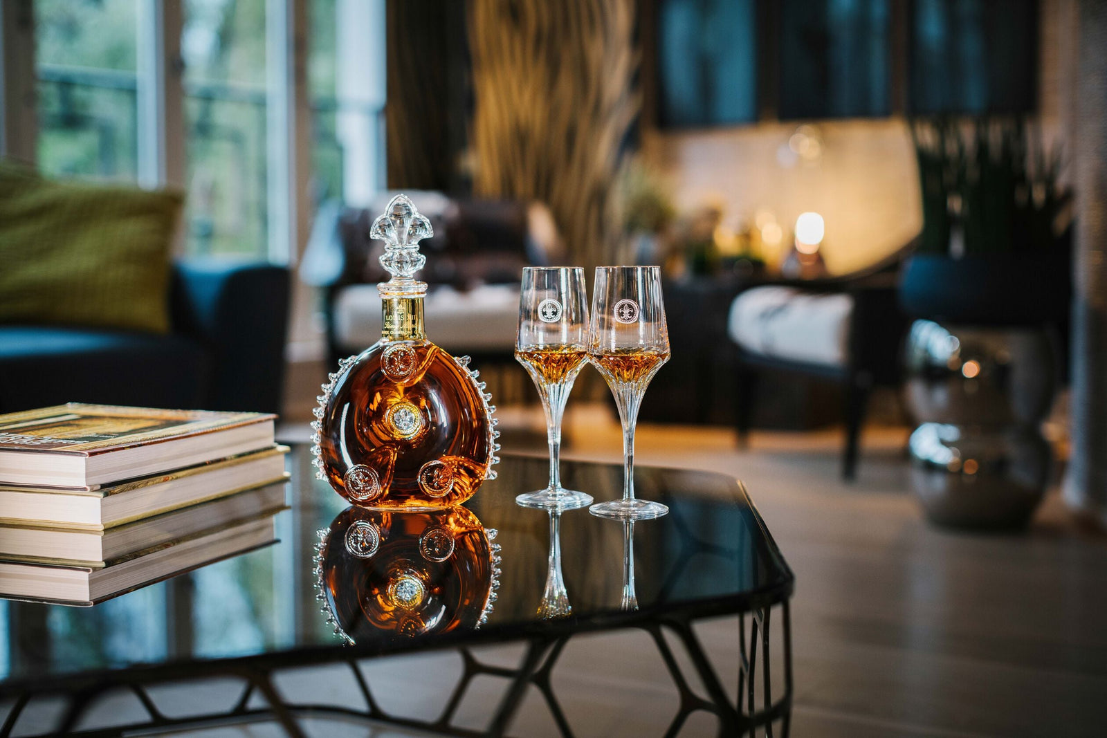 A lifestyle photo of a LOUIS XIII decanter with two semi-filled crystal glasses on a piano, a pile of books close by