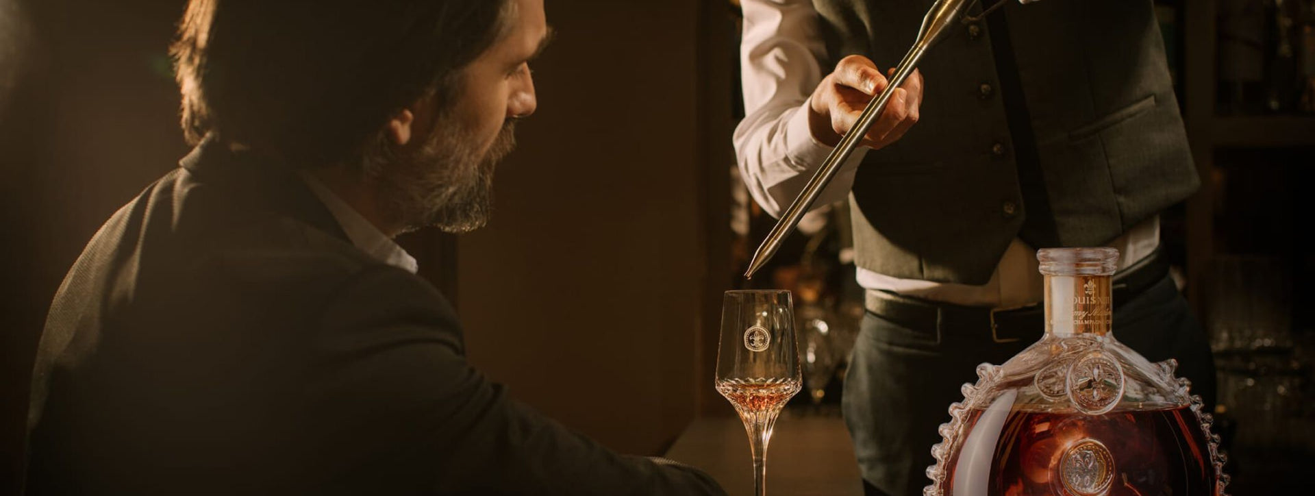 A photo of a man watching a bartender serving LOUIS XIII to a crystal glass with a spear, decanter nearby