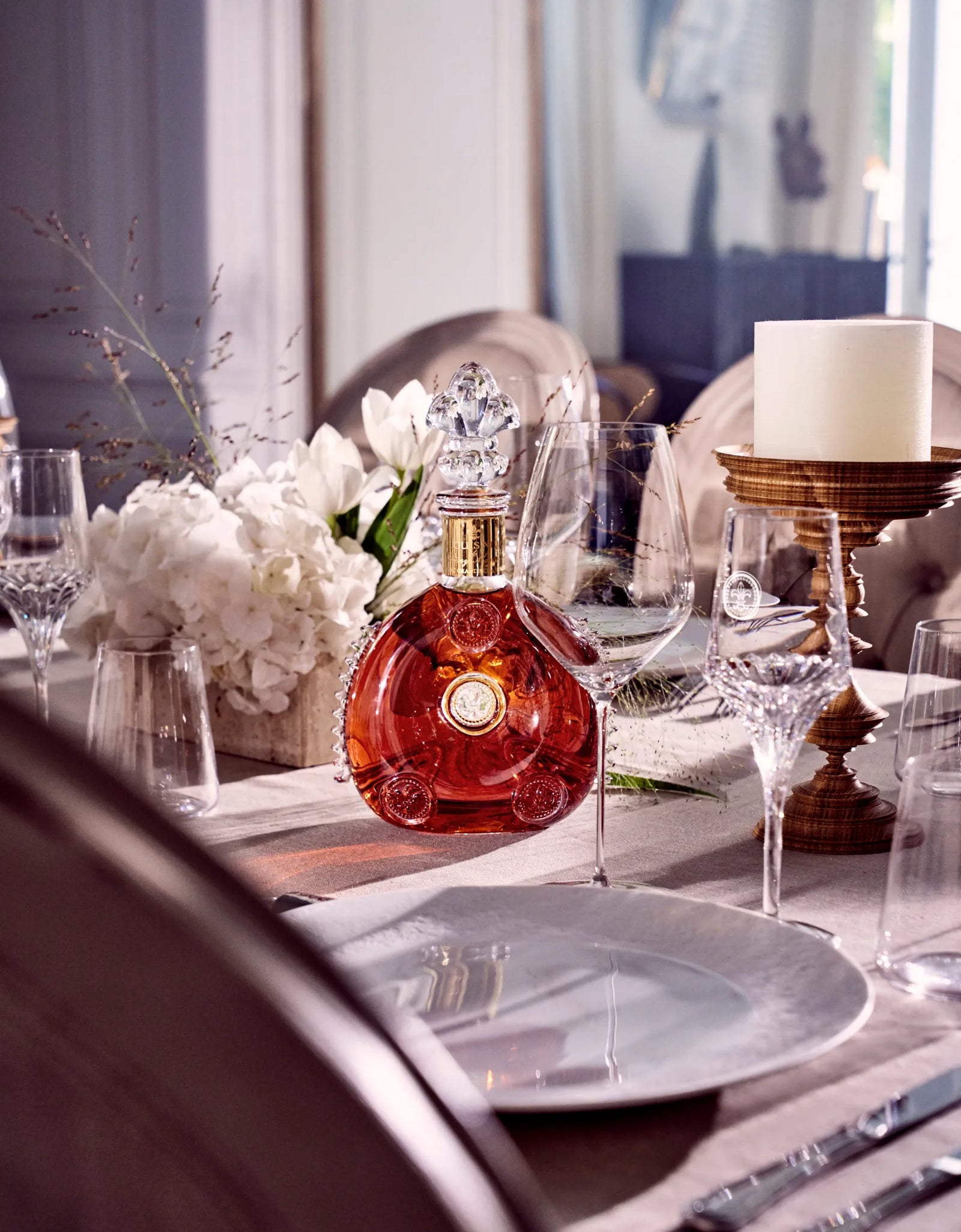 Bottle of Louis XIII cognac placed among crystal glasses, a candle on a wooden stand and a bouquet of flowers on a festive arrangement