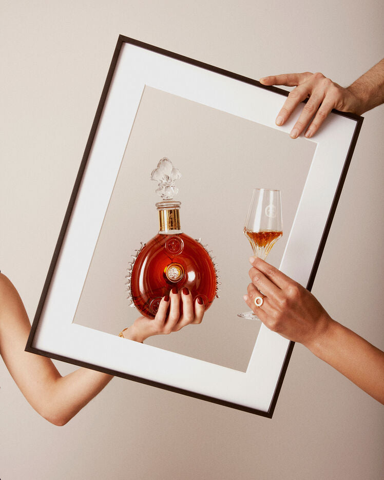 A lifestyle photo of two hands, one holding LOUIS XIII decanter, other a crystal glass, another hand holds an image frame framing decanter and the glass