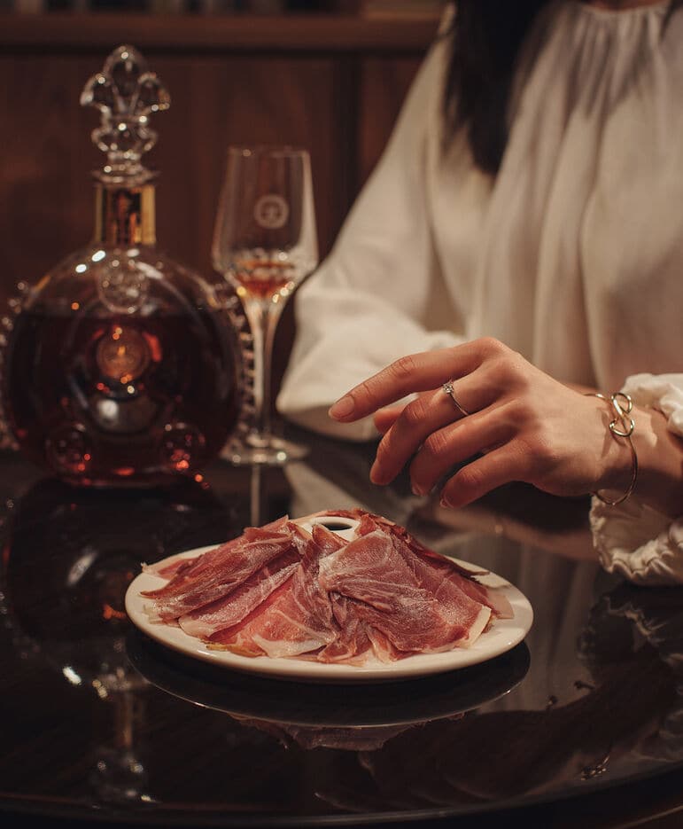 A photo of a female hand picking bellota ham from a plate,  a LOUIS XIII decanter with one crystal glass in the background