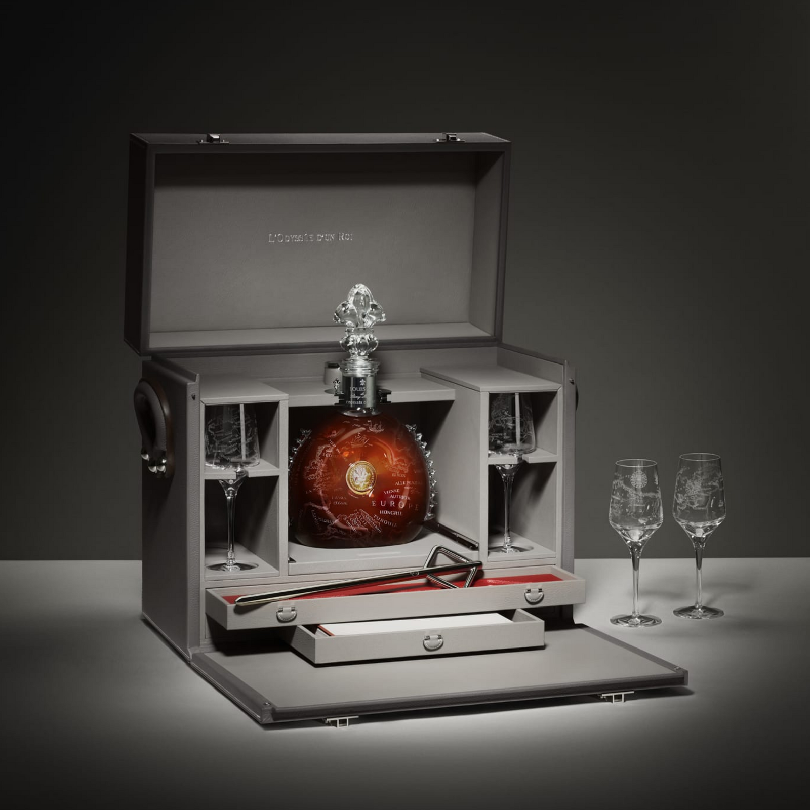 A lifestyle photo of LOUIS XIII L'Odyssee d'un Roi decanter in its box with two cristal glases in the box, a spear on the box drawer and two other crystal glasses nearby. Grey background